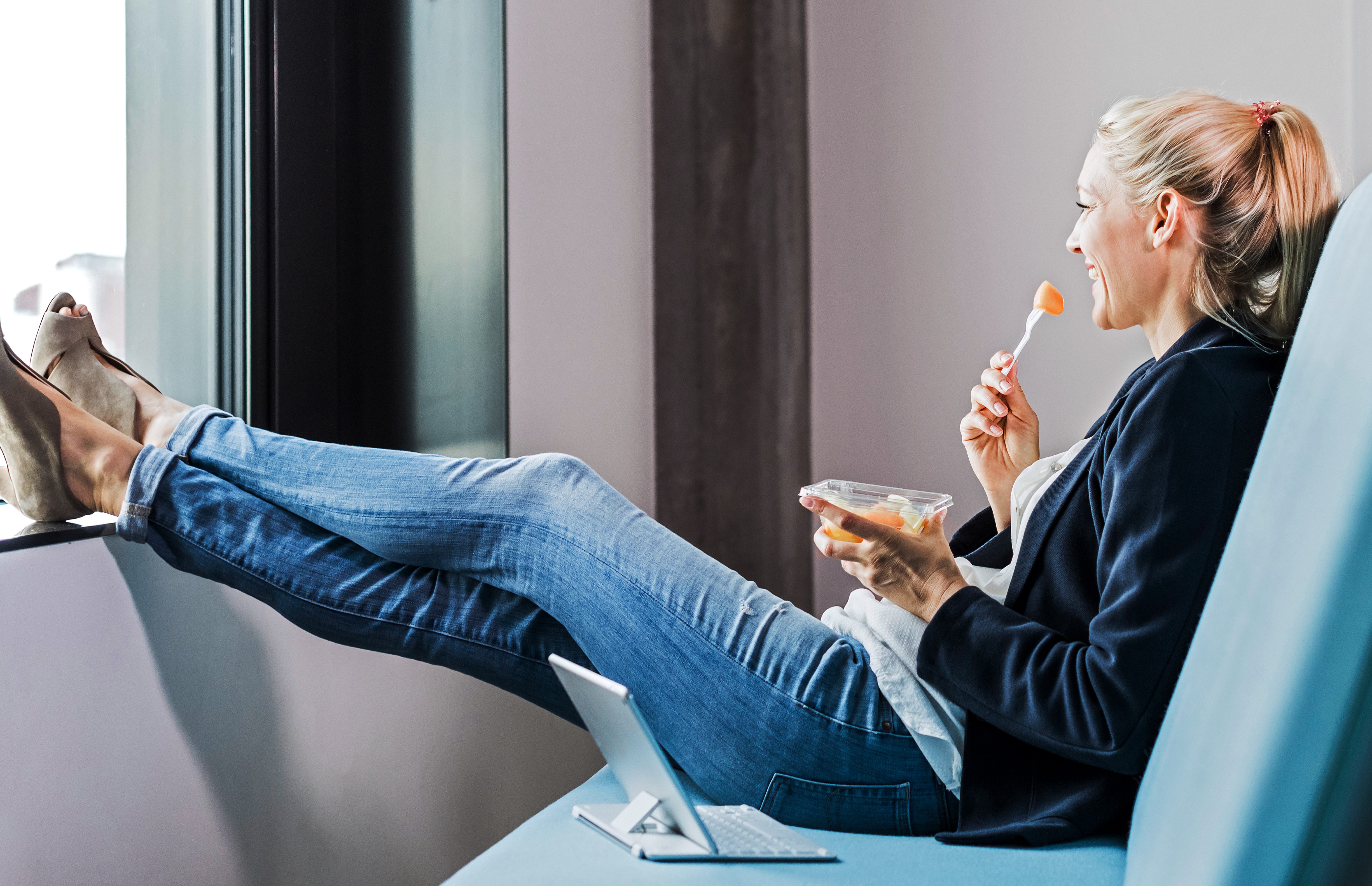 Eine Frau sitzt entspannt auf einem Sofa. Sie hlt eine Schale mit Essen in der Hand und isst mit einer Gabel. Vor ihr steht ein aufgeklappter Laptop.