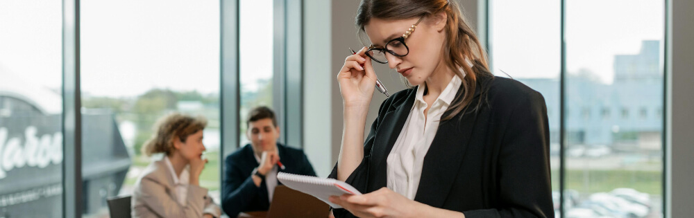 Eine Geschftsfrau in einem dunklen Anzug und mit Brille steht in einem modernen Brogebude und sieht auf ein Notizbuch in ihrer Hand. Im Hintergrund sind zwei Personen in Businesskleidung zu sehen, die an einem Tisch sitzen und miteinander sprechen. 